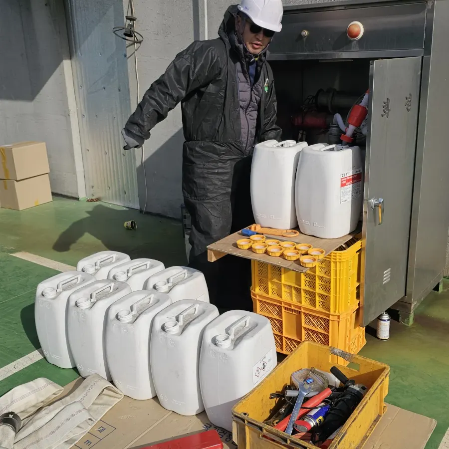 안전모를 쓴 작업자가 포소화전 캐비닛 앞에서 약제 용기를 다루고 바닥에 흰색 약제 용기가 줄지어 놓인 모습