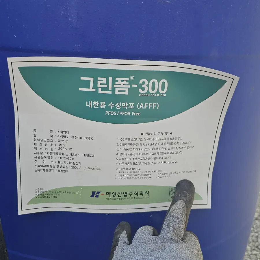 청색 드럼에 붙은 그린폼-300 내한용 수성막포 AFFF 라벨, 취급 설명 부분을 가리키는 장갑 낀 손